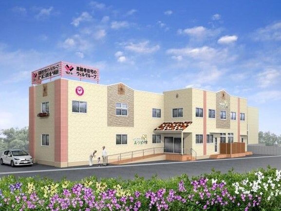 草花に囲まれた２階建ての建物はベージュ系で玄関には手すり付きのスロープと屋根があり、明るい雰囲気。