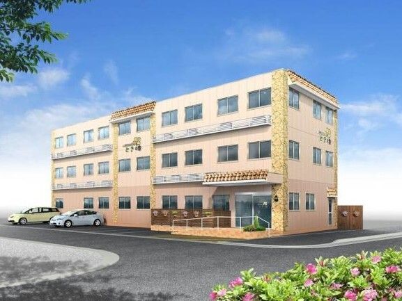 ３階建ての建物はベージュ系で敷地内には駐車場があり、車が止まっている。玄関には手すり付きのスロープが付いている。