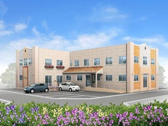 ベージュ系の２階建ての建物。玄関には手すり付きのスロープが付き、屋根も設置されている。
