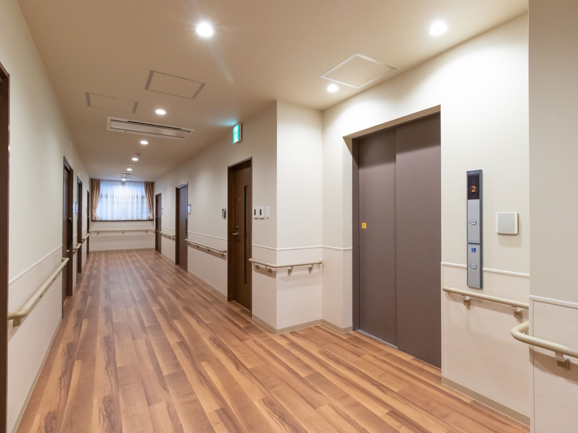 エレベーター前に伸びた木目のきれいな床の廊下である。突き当りの高窓からは自然光が入っている。廊下の両側壁には手すりが設置されている。