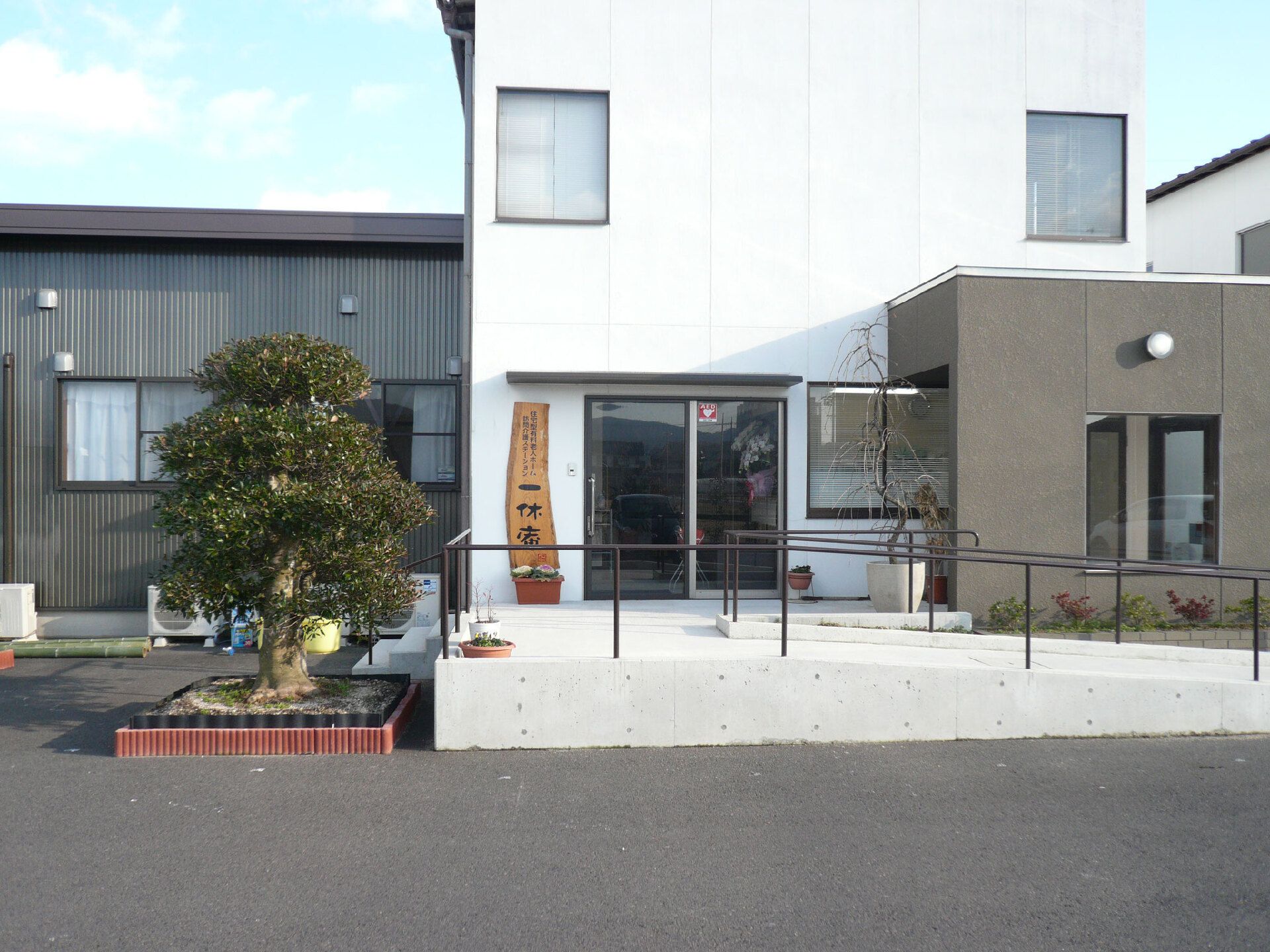 2階建ての白とグレーでまとめられた建物の玄関には手すり付きのスロープがあり、木も植えられている。