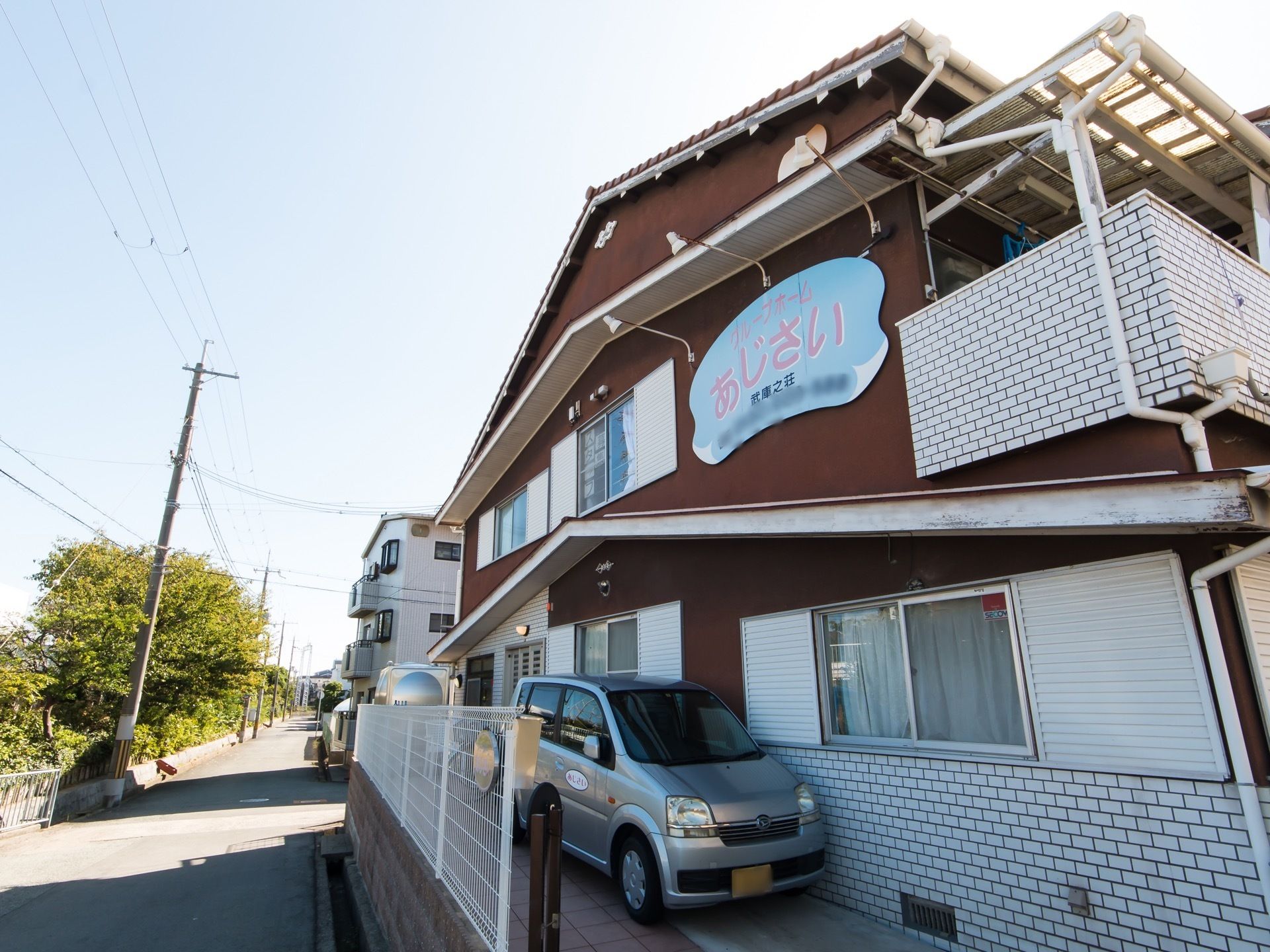 ２階建て施設の壁面には、施設看板がある。駐車スペースが壁沿いに設けられ、車が１台停車している。