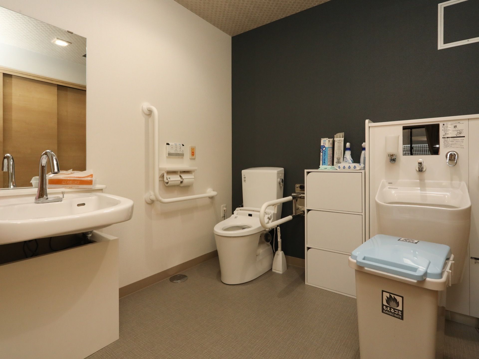 広々としたトイレである。手すりやオストメイト専用洗面台、呼び出しボタンが完備されている。自動水栓付き洗面台も設置されている。