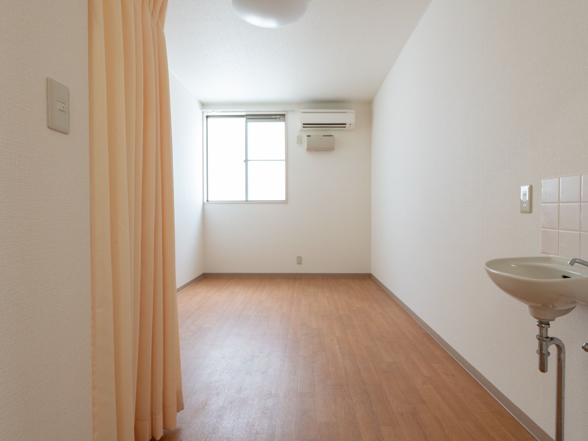 壁は白色で、床はフローリングの個室になっている。右側に洗面台が設置されている。窓の近くにエアコンが備えつけられている。