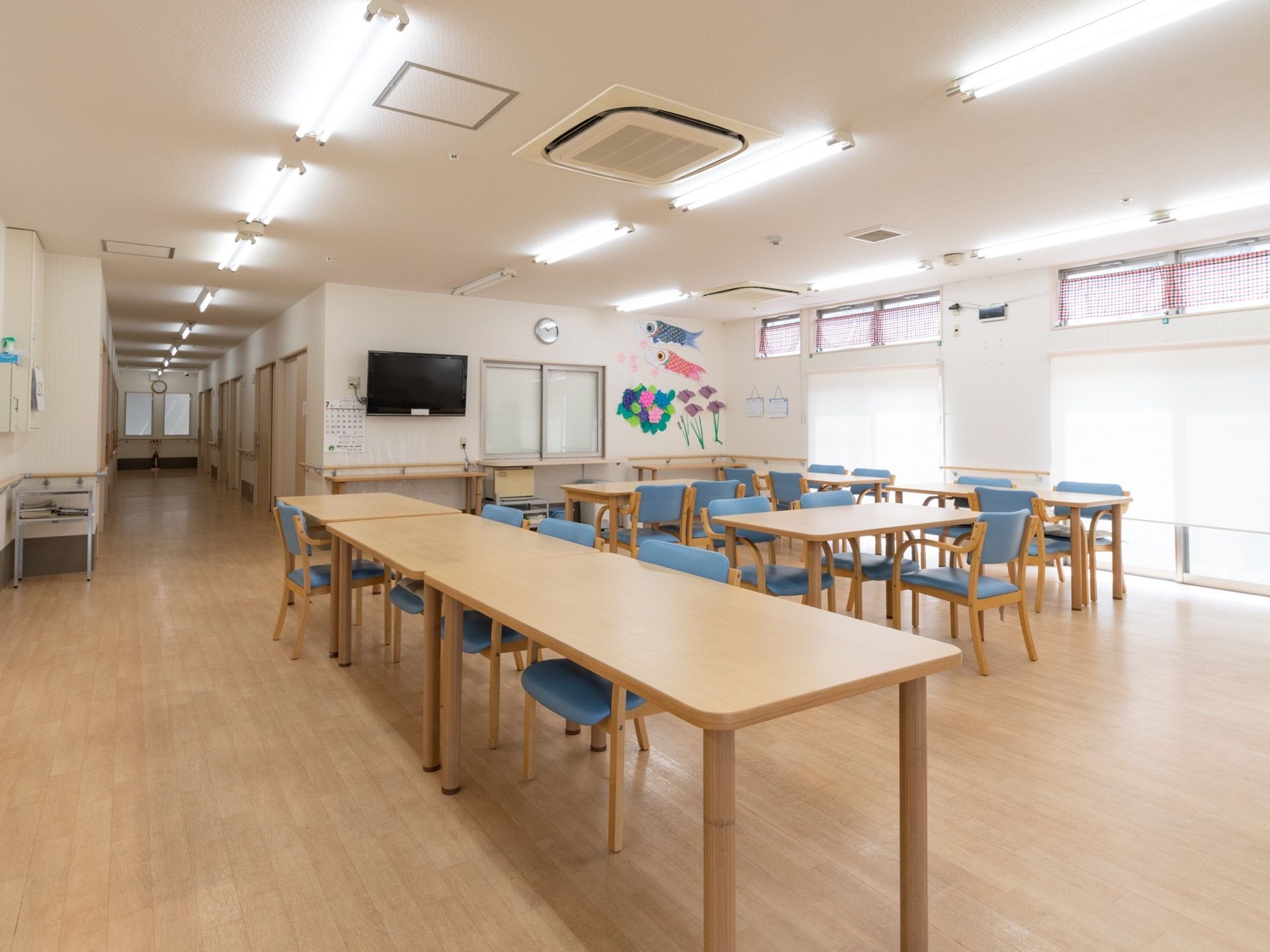 木製の長テーブルと青いクッションがついた椅子が複数置かれている。壁にテレビが掛けられており、周囲には折り紙で作られた作品が飾られている。