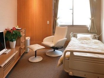 カーペットが敷かれた居室にはベッドやテレビなどの家具家電があり、オットマン付きの椅子も置かれている。