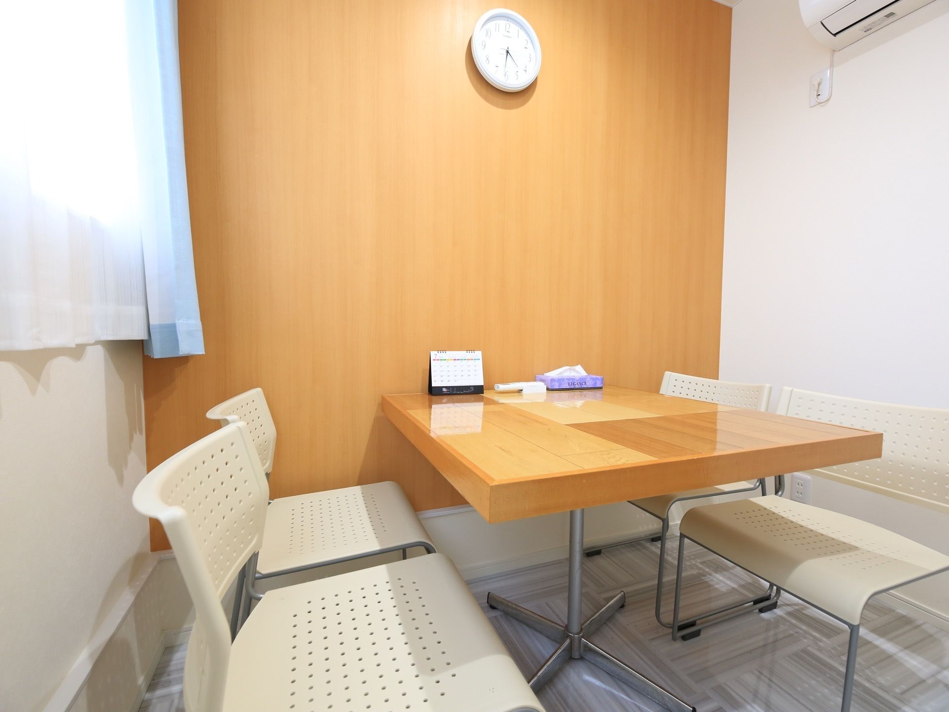 木製のテーブルと白い椅子が４脚置かれている。テーブルの上には、卓上カレンダーとティッシュがあり、壁には時計がかけられている。