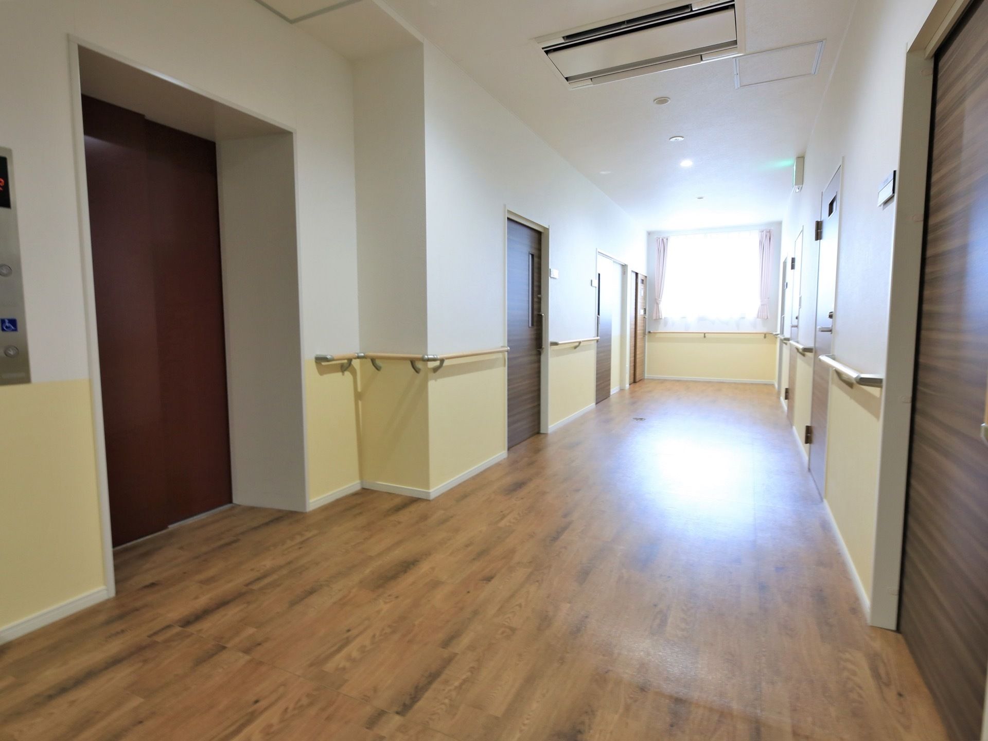 フローリングの床の廊下である。壁が２色で構成されており、手すりが取り付けられている。また、廊下のつきあたりには、窓が設置されている。