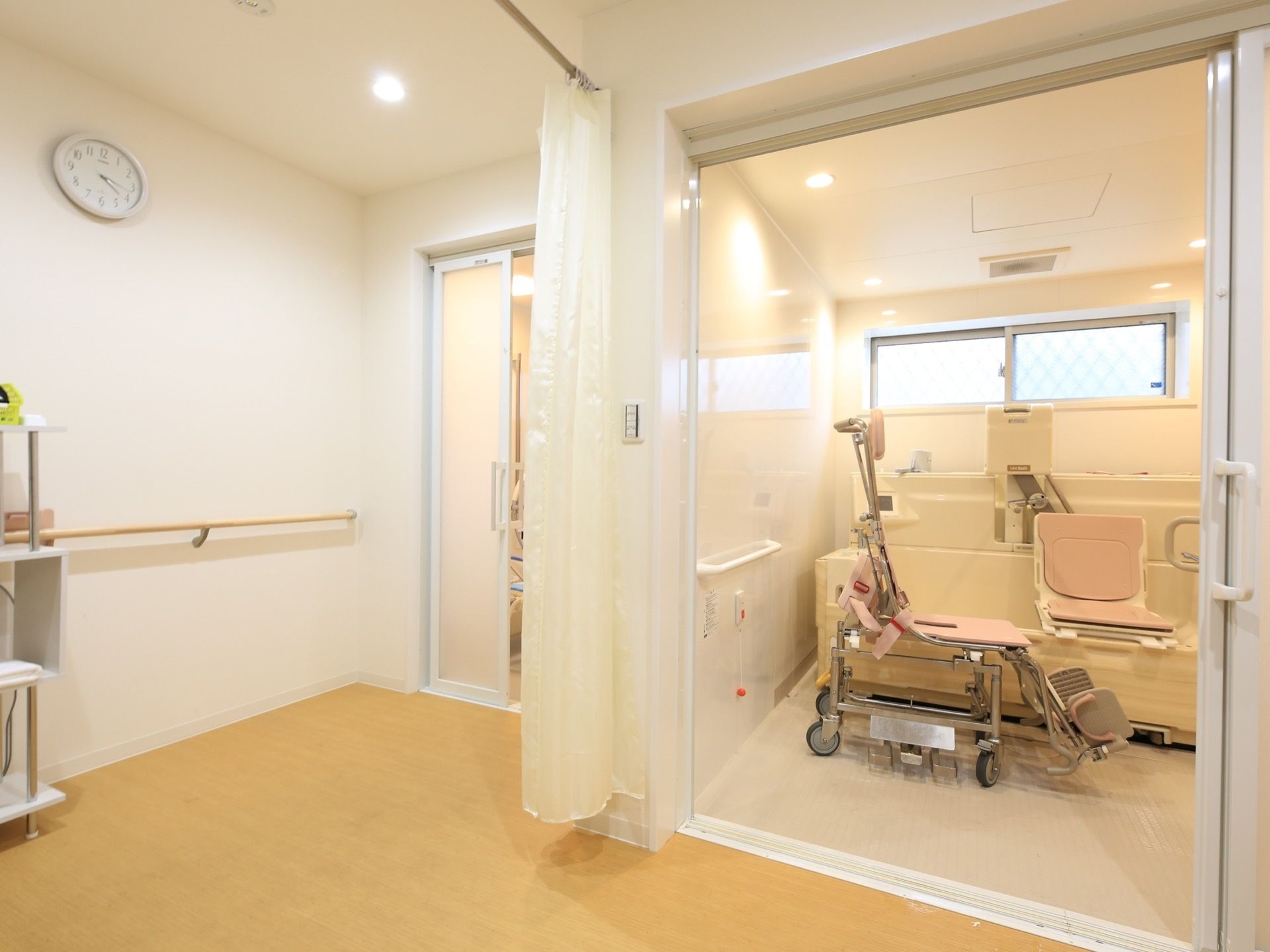 白い壁には時計があり、手すりが取り付けられている。浴室は２箇所あり、その間に仕切り用のカーテンがかかっている。
