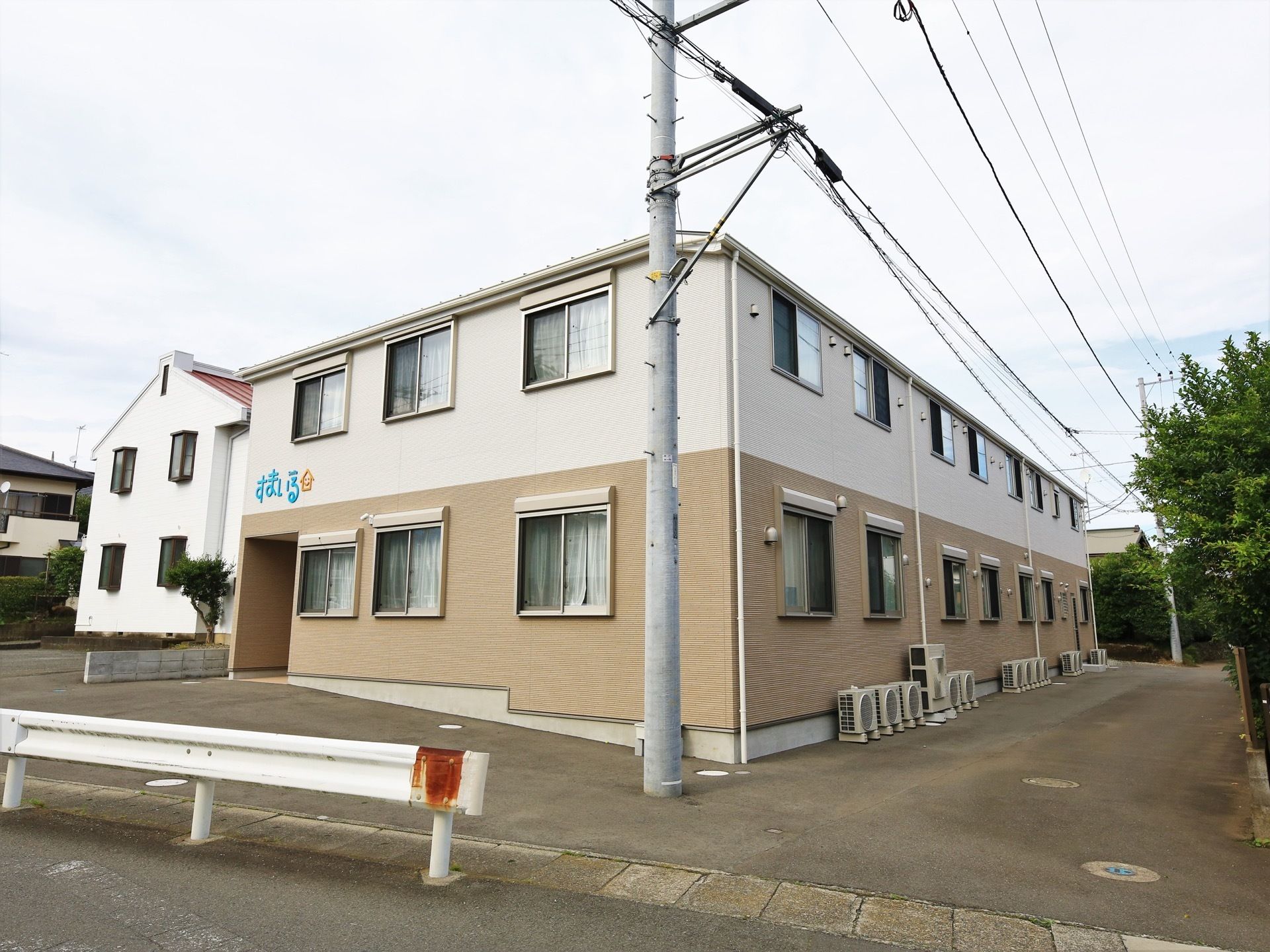 ベージュと白の２色で構成された２階建ての建物である。窓の下にはエアコンの室外機が並べて置かれている。
