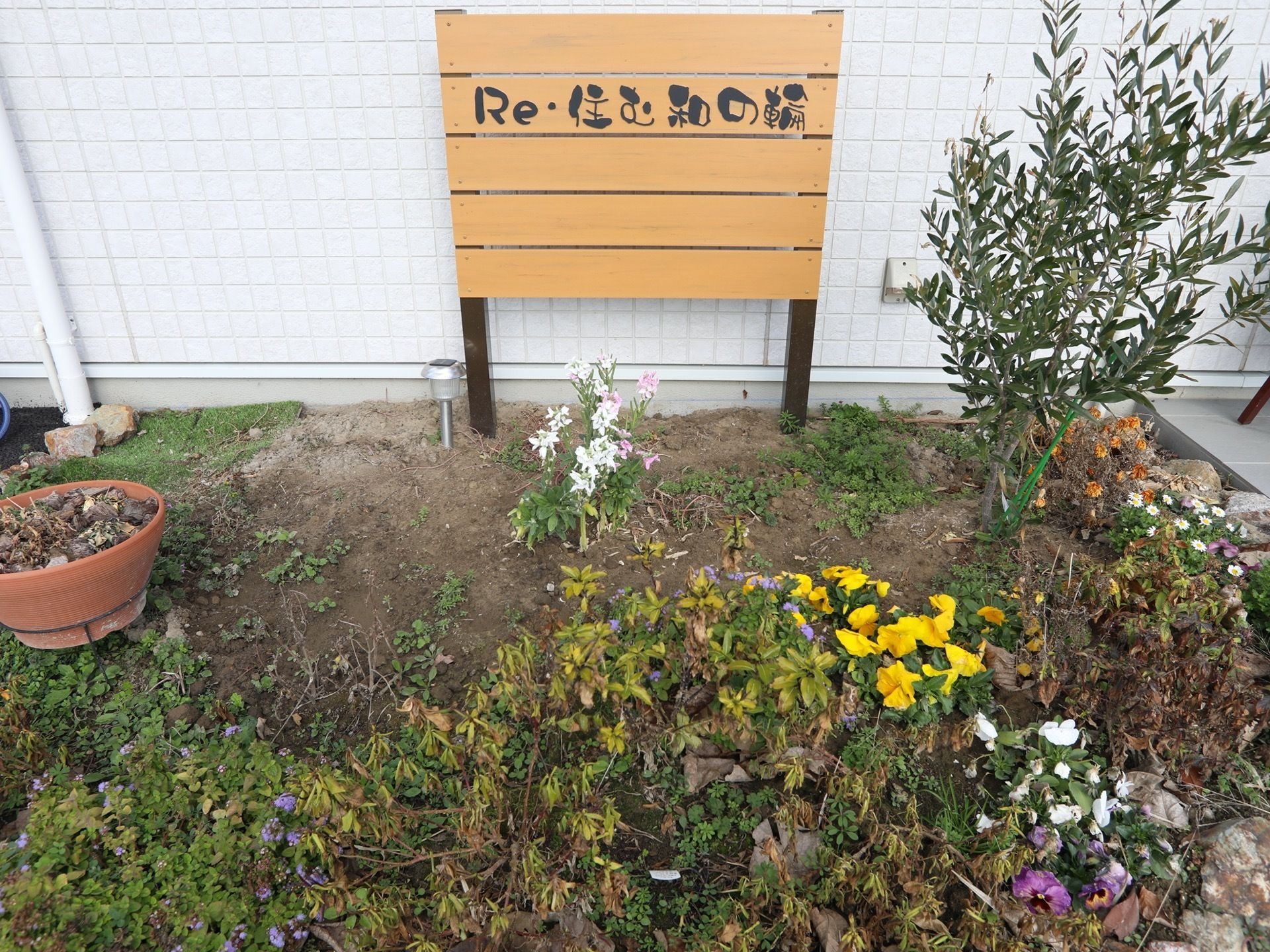 小さなかわいらしい雰囲気の花壇が設けられている。数種類の花が植えられており、奥には施設名の書かれた木目調の看板が立てられている。