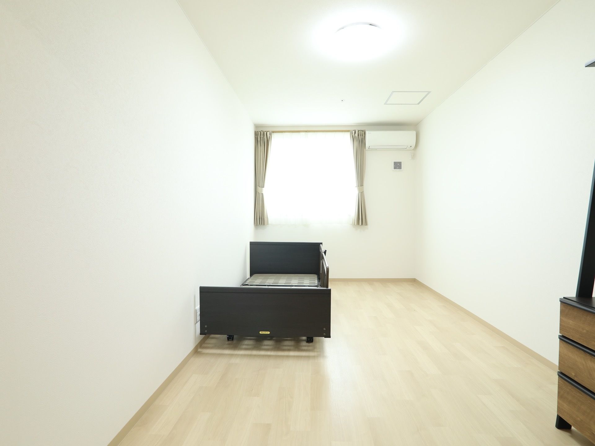 色を基調とした明るいフローリングの居室である。奥には腰高窓があり、壁の上の方にはエアコンが設置されている。