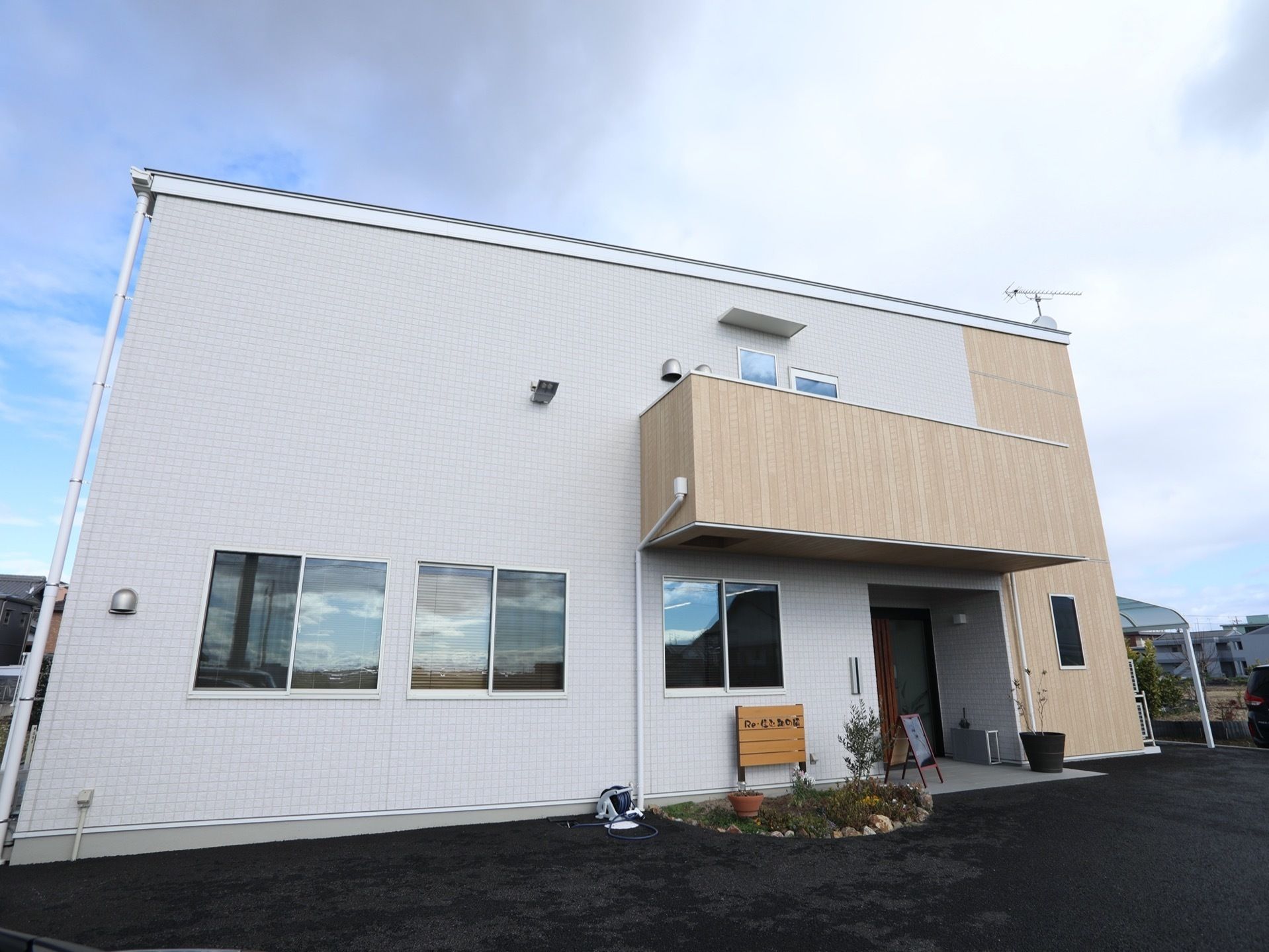 建物は全体的に白い外壁で、右側の一部と玄関のひさし部分は木目調になっている。玄関の側には小さな花壇があり、横にホースが置かれている。