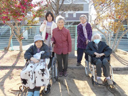 芝生の上で座って笑顔で話をしている高齢夫婦と女性。女性二人は白いシャツを着ている。