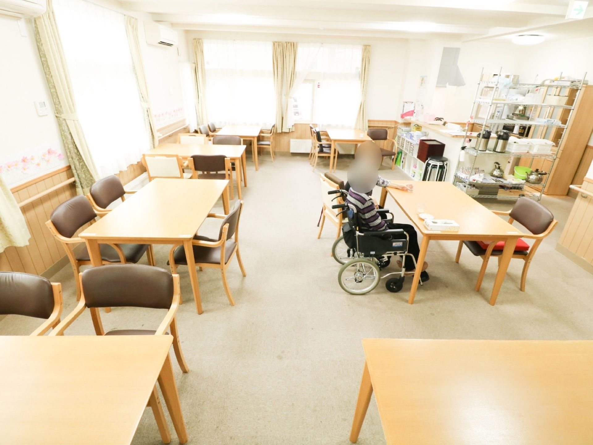 食堂に２名の入居者様がいる。一人は車椅子に、もう一人は食堂の椅子に座っている。テーブルの上にティッシュペーパーの箱が置かれている。
