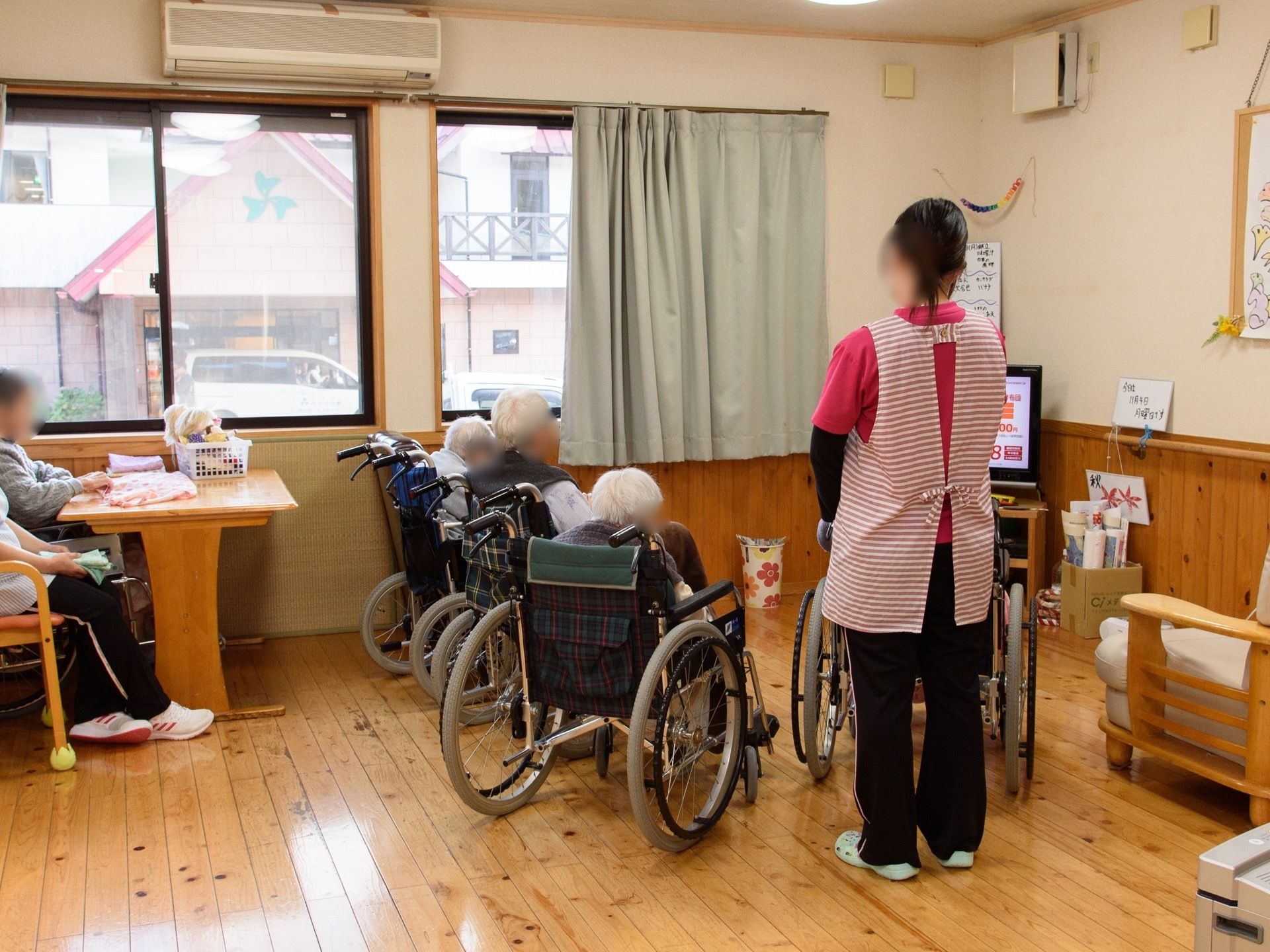 共有スペースに多くの入居者様が集まっている。車椅子をご利用の方が数人でテレビを囲んでいて、その後ろに女性スタッフが立っている。