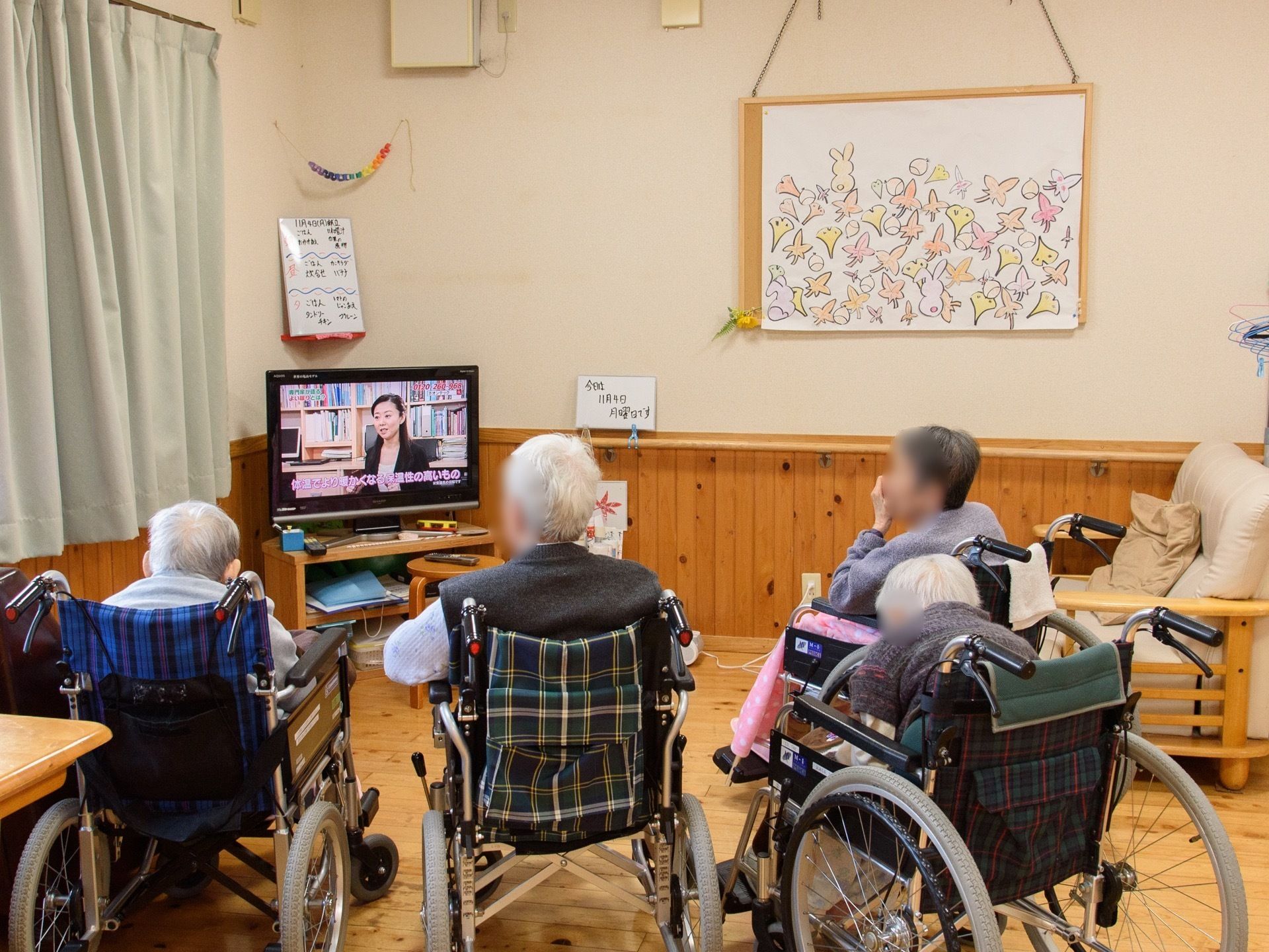 共有スペースにあるテレビの周りに、車椅子をご利用の入居者様が４名集まっている。壁際にはソファが置かれている。