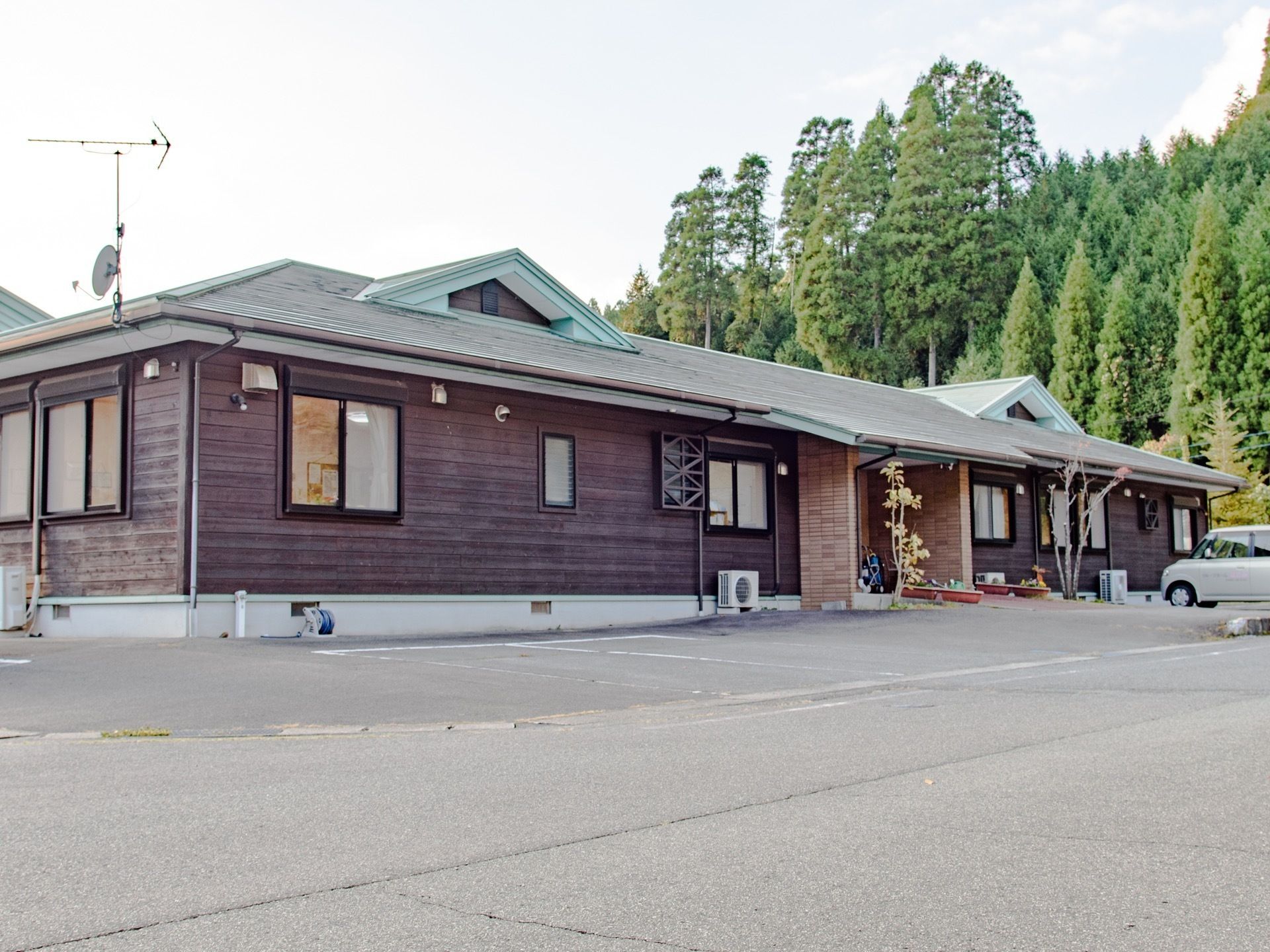 後ろに大きな木々が生えている敷地に建つ平屋建ての施設である。壁は木の板で、玄関前に車が１台停まっている。