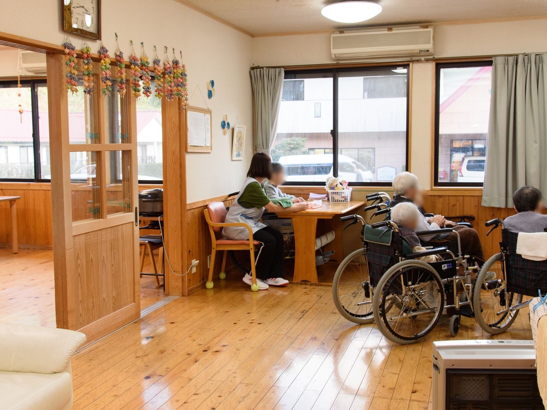 共有スペースの窓際にあるテーブルに、入居者様と女性スタッフが並んで座っている。その窓の上にはエアコンを設置。