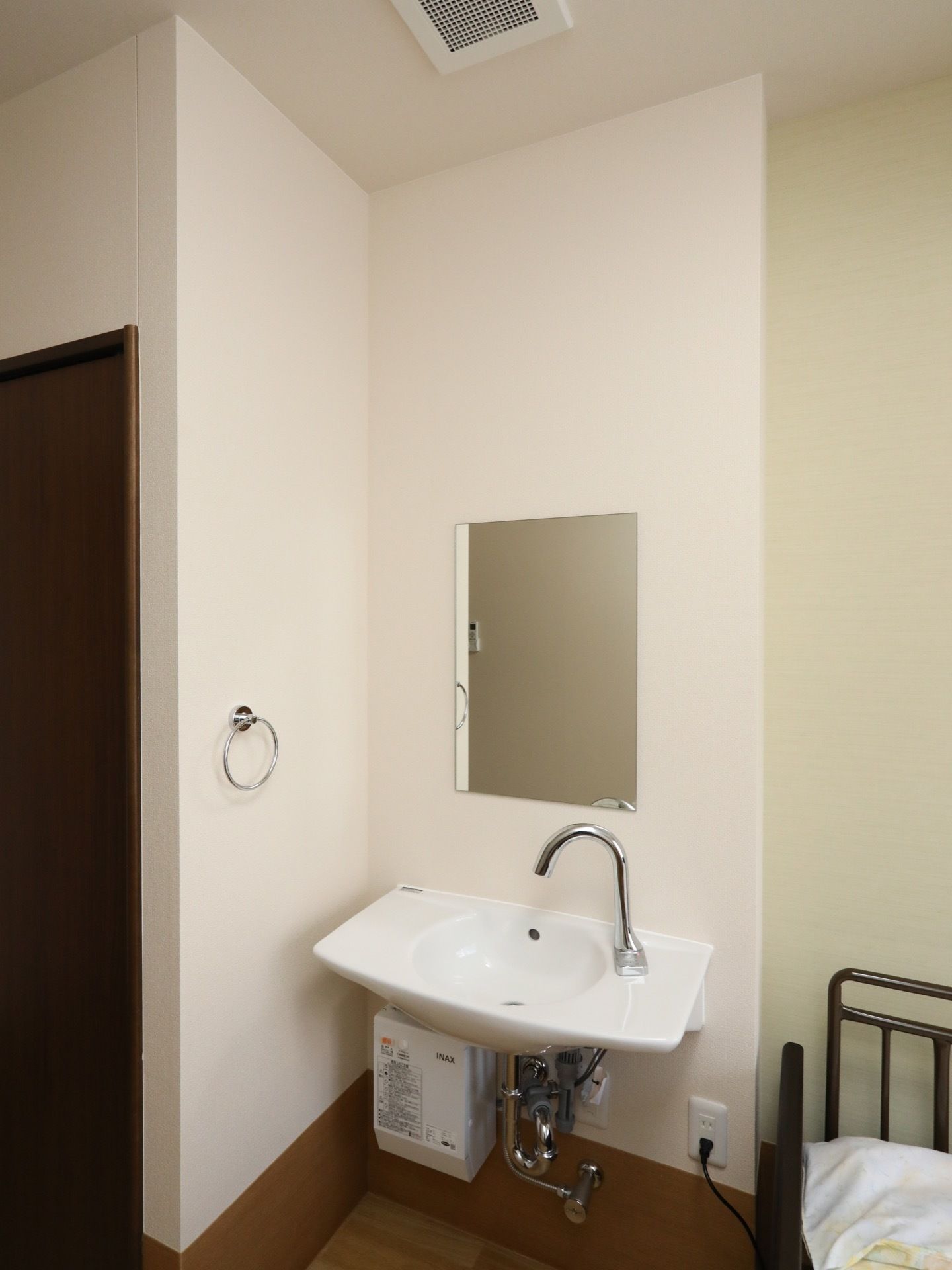 給湯器が設置された洗面台があり、正面の壁に鏡がかかっている。布団が敷かれたパイプベッドの角が見えている。