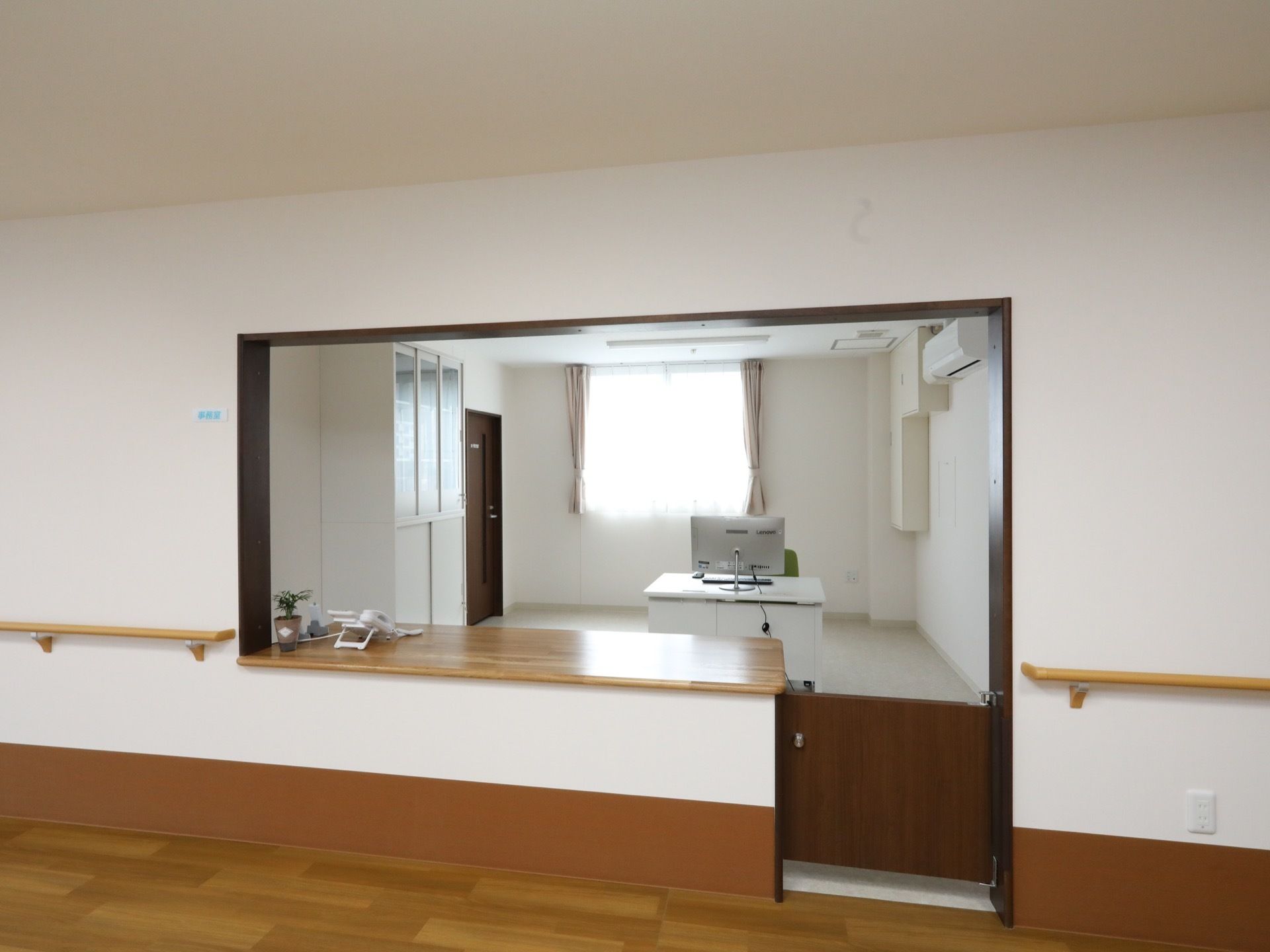 木目の天板のカウンターがあり、横に小さな扉がある。カウンターの奥の部屋にある白いデスクにパソコンが置いてある。