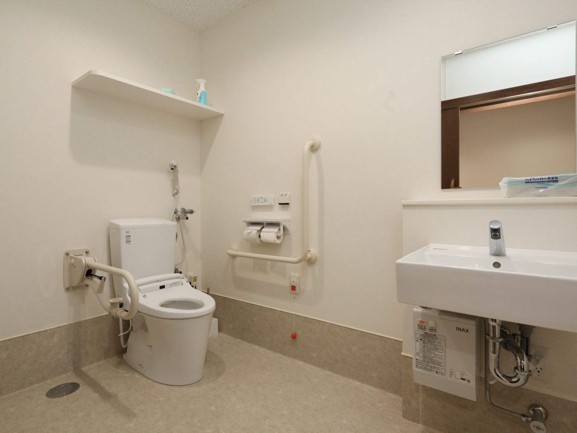 白いタンクの洋式トイレがあり、両側に手すりがある。湯沸かし器が付いた白い洗面台があり、棚の上にペーパータオルが置いてある。