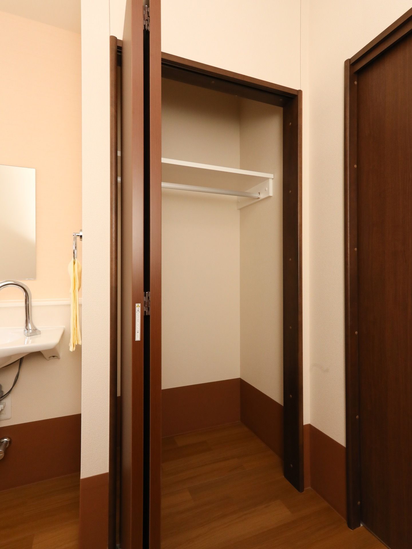木目のフローリングの床の部屋にクローゼットと洗面台がある。クローゼットの扉が開かれ、中に棚がある。