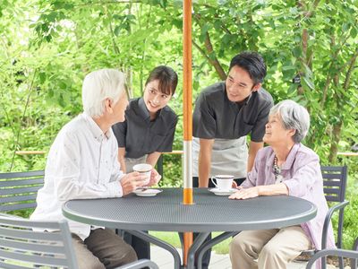 緑あふれる庭園でティータイムを楽しむ高齢の男女と、その二人に笑顔で話かける男女のスタッフ。