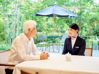 白髪の男性とスカーフをした女性スタッフが話をしている様子。テーブルにはテーブルクロスがかけられ、窓の外には庭がある。
