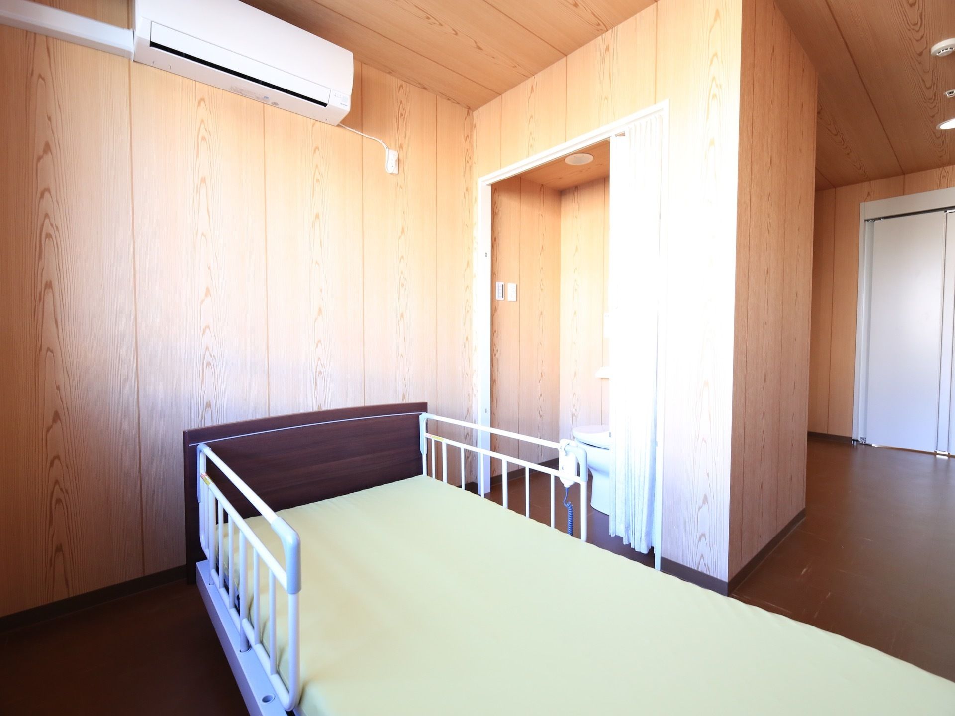 居室の天井付近には、エアコンが取り付けられている。ベッドの右横にはトイレがあり、仕切りカーテンが端に寄せて開けてある。