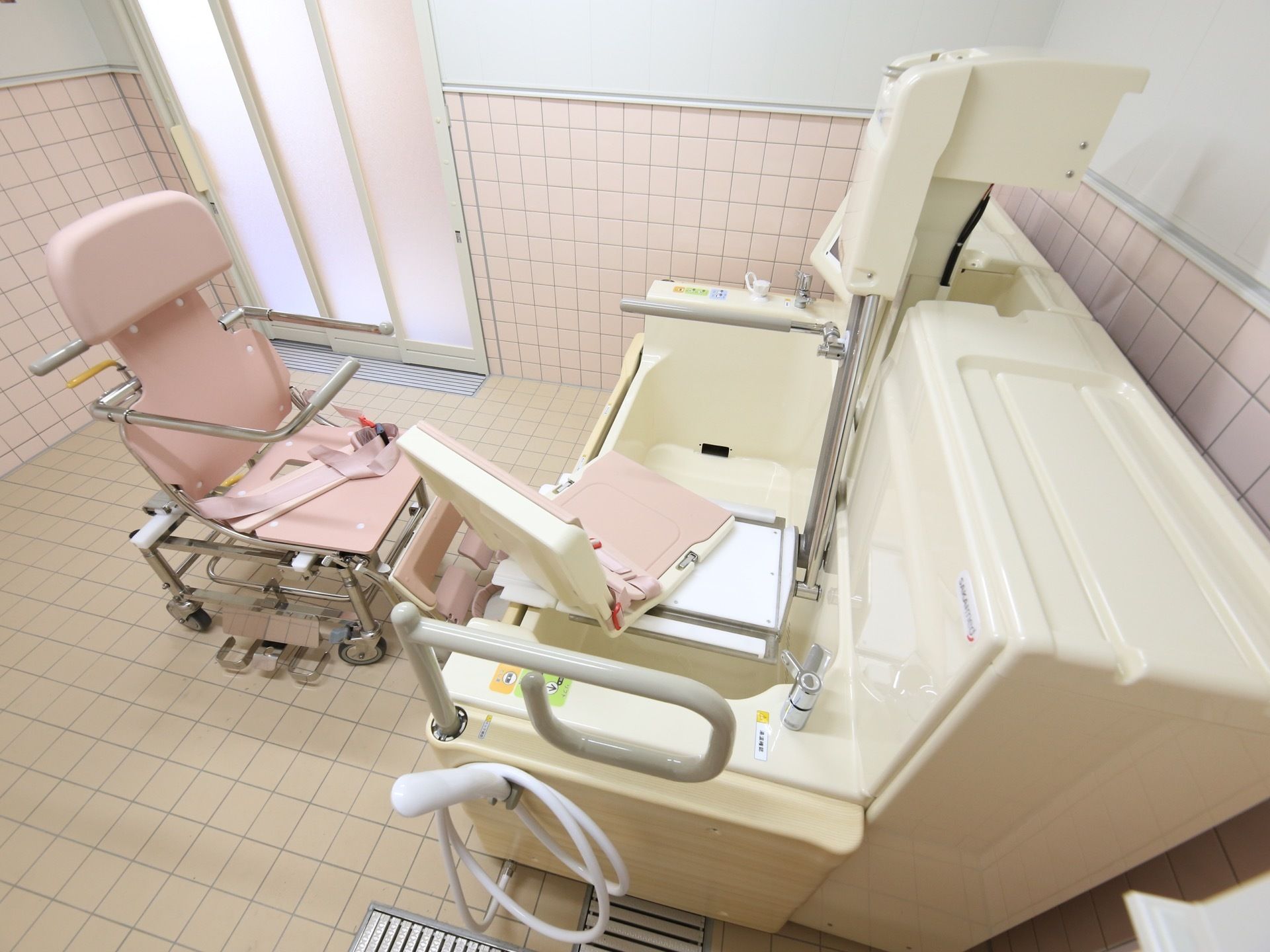 大きな介護用入浴装置が、浴室の左側に置いてある。壁に貼り付けられたタイルは淡いピンク色で、椅子と同系色になっている。