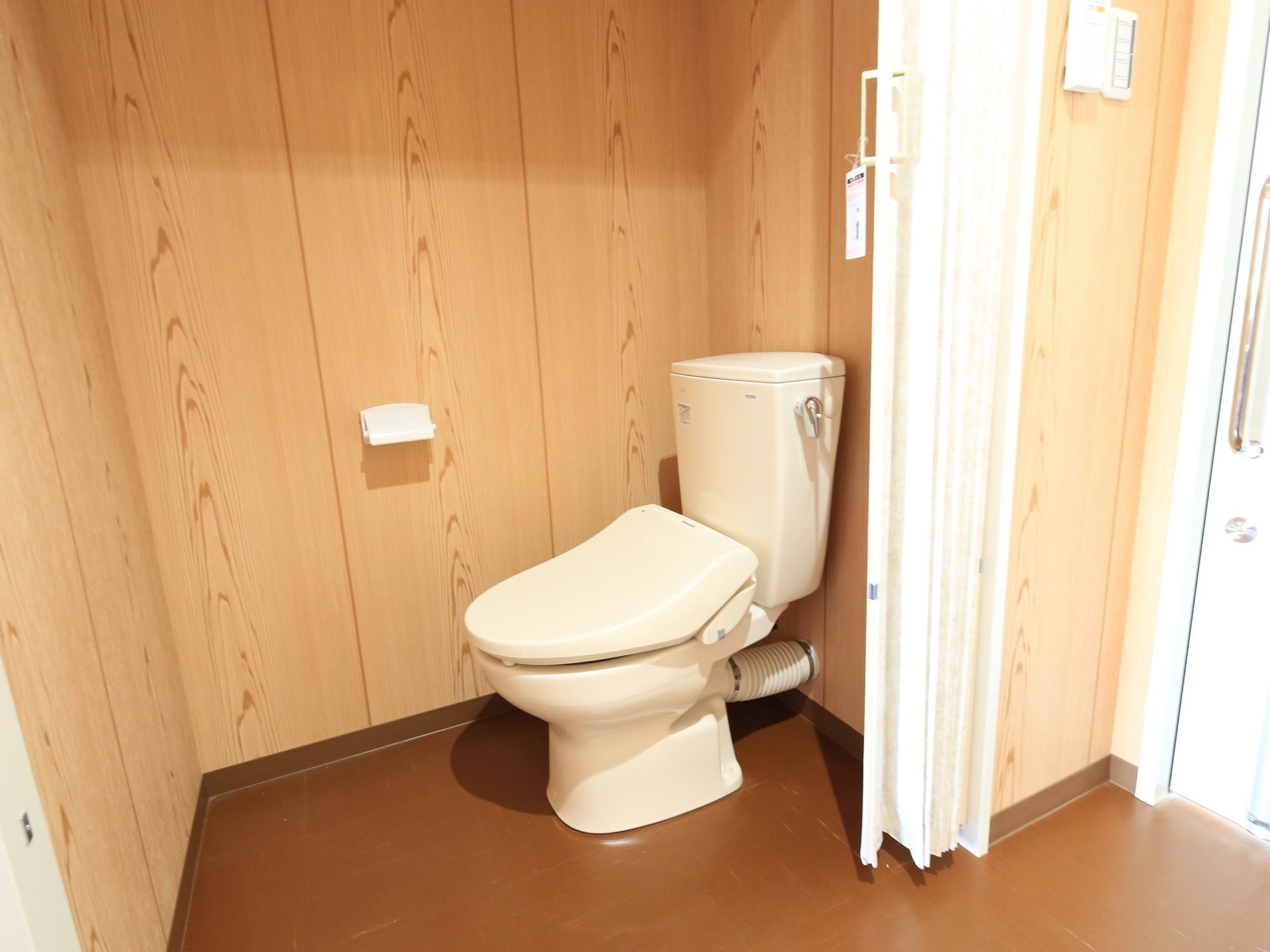 白とブラウン系の色でまとめられた落ち着いたデザインのトイレである。ドアの代わりに、アコーディオンカーテンが付いている。