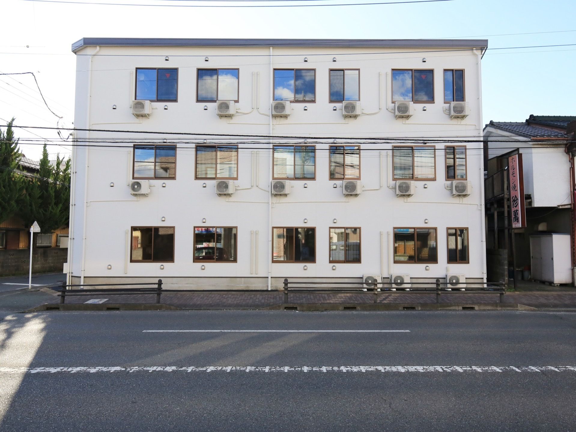 白色で統一された３階建ての施設である。窓が並び、それぞれエアコンの室外機が設置されている。建物の横に歩道があり、その隣には飲食店がある。