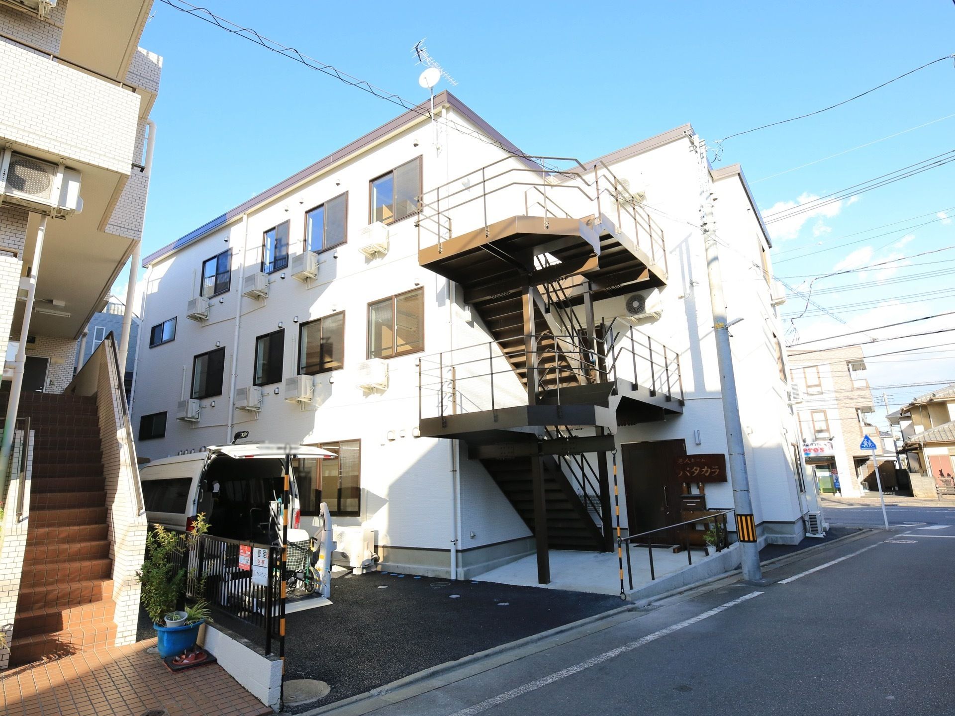 公道に囲まれた角地に、白い外壁の３階建ての施設が建っている。建物の左側のスペースがアスファルトで舗装されており、福祉車両が駐車してある。