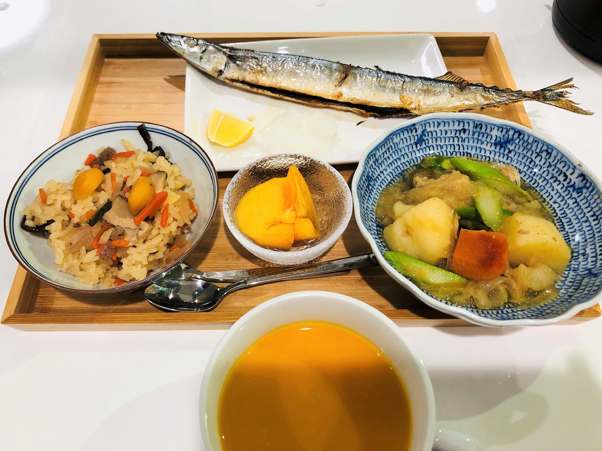 木製のトレーの上にはサンマや炊き込みご飯、煮物などが置かれ、手前にはスープが入ったコップがある。
