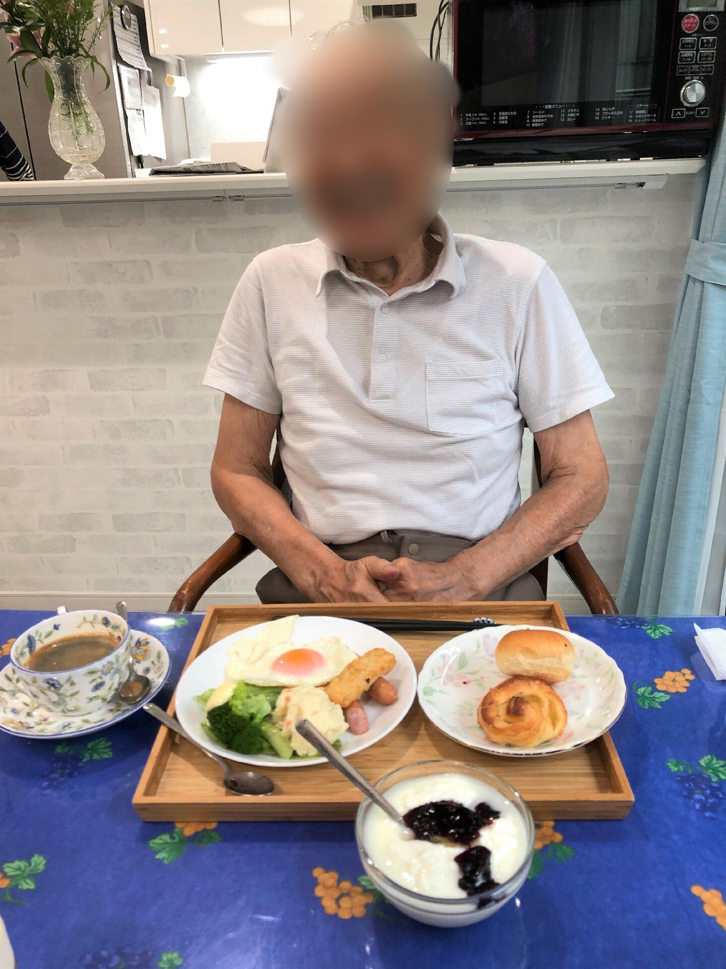 木製のトレーの上にはパンが二つ置かれた皿とソーセージや目玉焼き、野菜が盛られた皿があり、トレーの外にはコーヒーやジャム入りのヨーグルトもある。