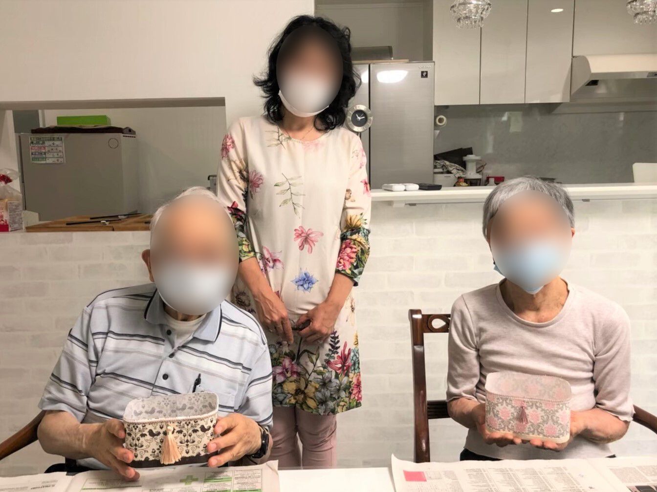 いすに座って作成した作品を披露している男女の入居者の後ろには女性が立っている。三人ともマスクをつけている。