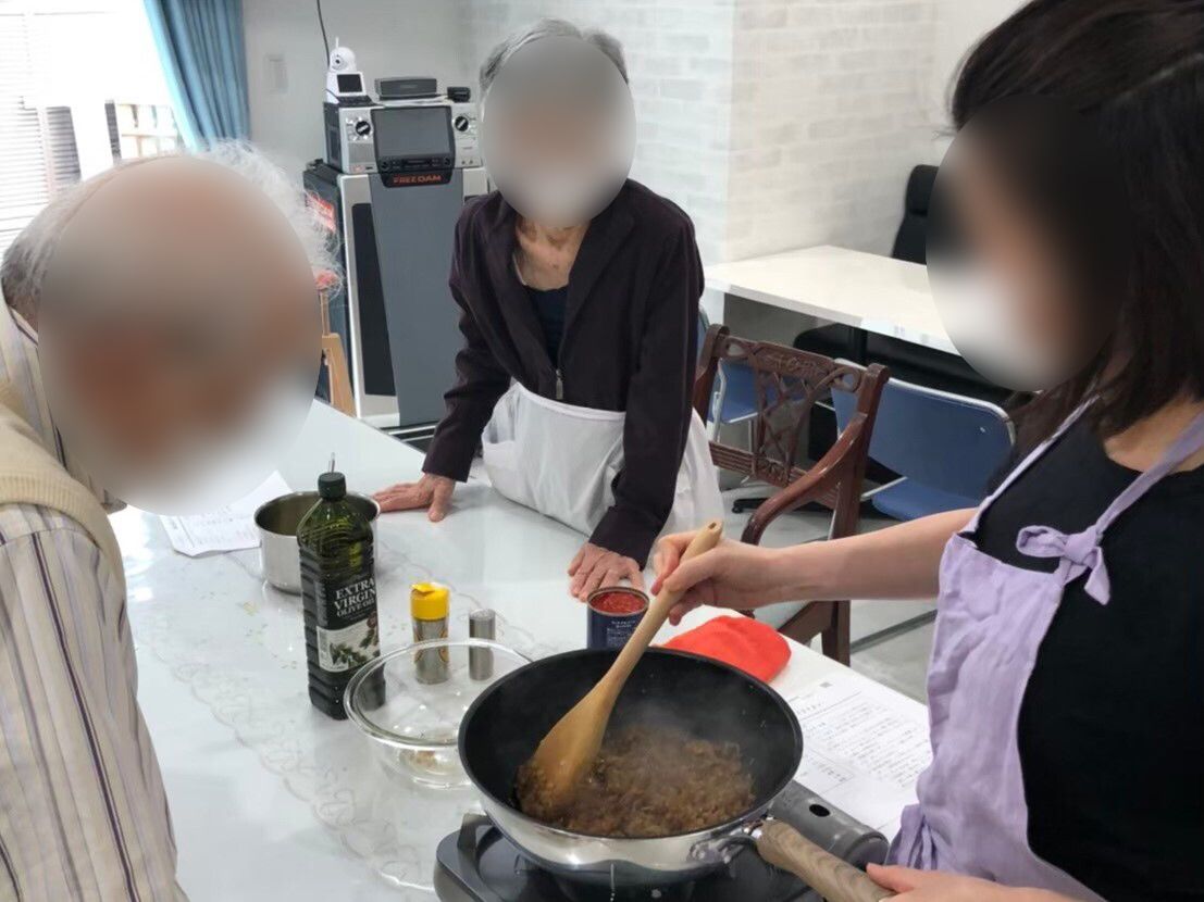 真剣な表情で女性講師の料理をする姿を見ている男女各1名の入居者。フライパンの中でひき肉のようなものを炒めている。
