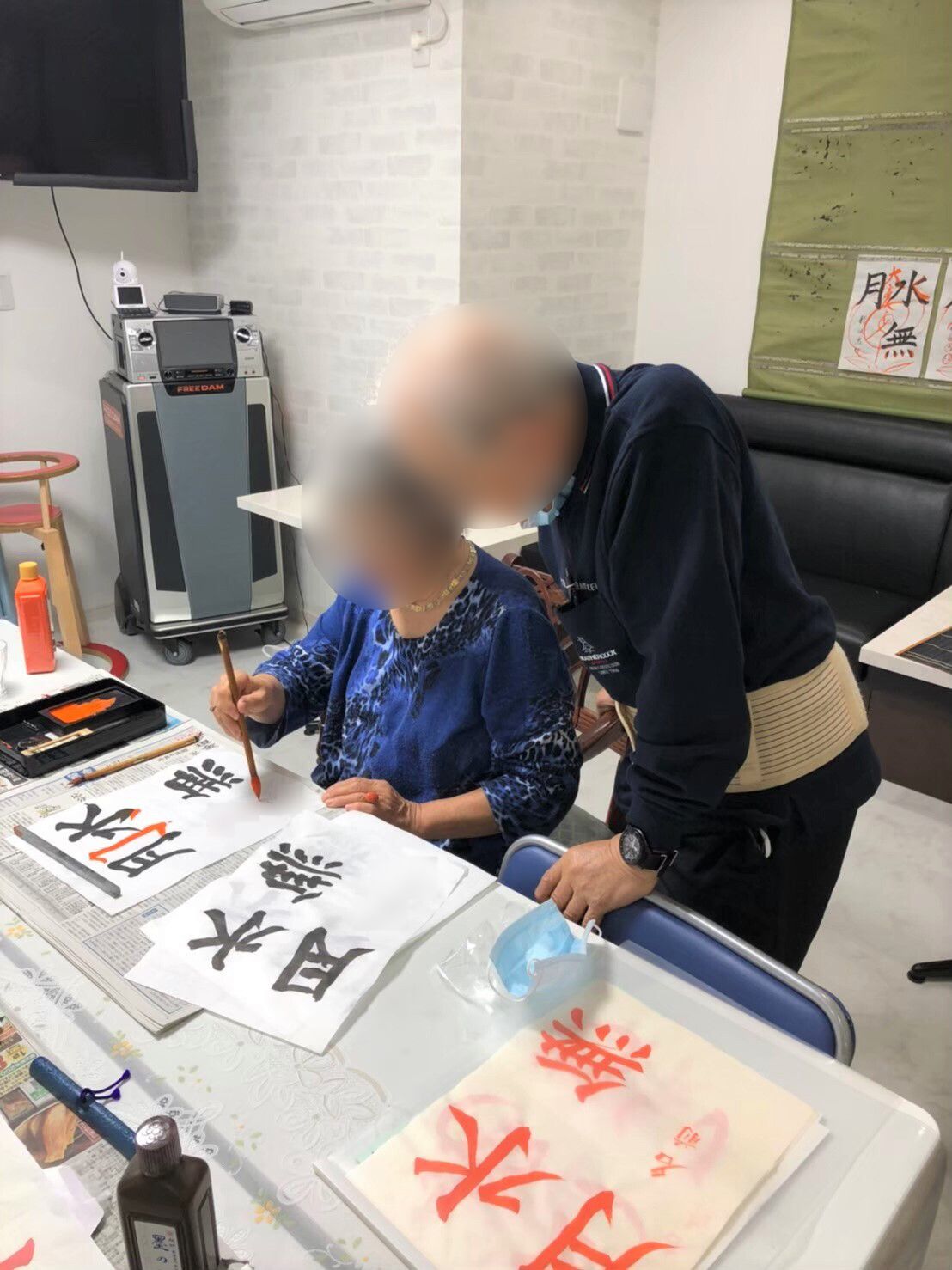 ブルーの服を着た白髪の女性が書道をしている様子。マスクをした高齢男性がその横で見ている。