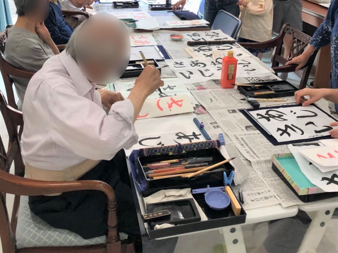 新聞紙が敷かれた長テーブルを囲み、男女の入居者が真剣に書道に取り組んでいる様子。「あじさい」という文字を書いている。