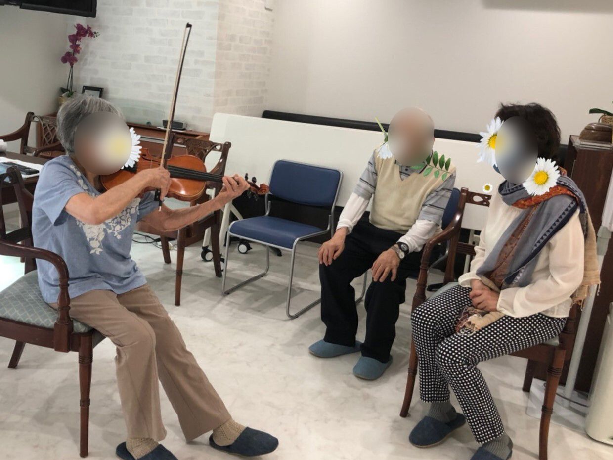 バイオリンを弾く女性入居者と、近くの椅子に座ってそれを聞いている男女の入居者。周りにも椅子が置かれている。