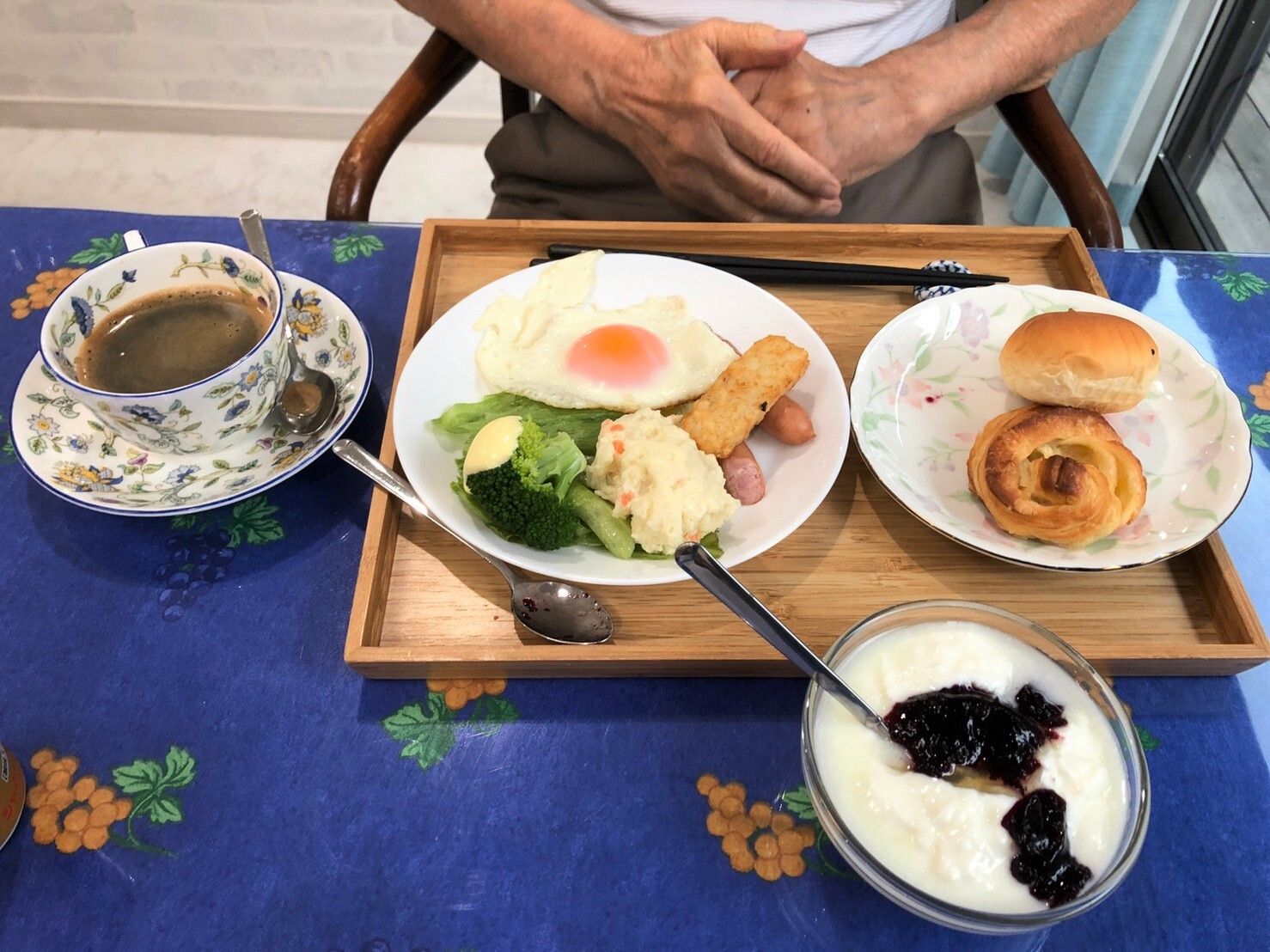目玉焼きとウインナー、野菜とポテトサラダが一つの皿に盛られ、隣にパンが２つ、ヨーグルトとコーヒーが用意された食事