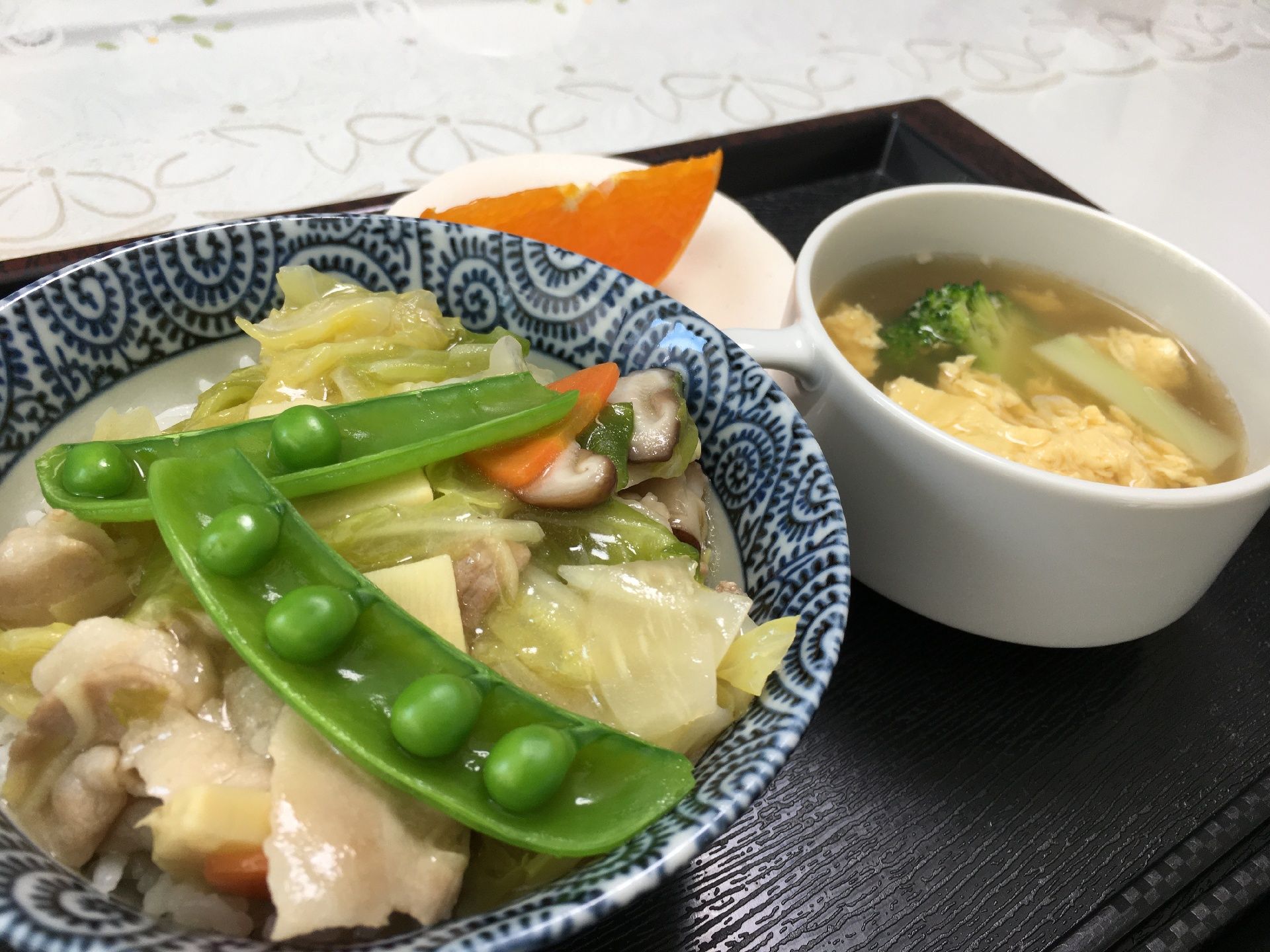 ご飯の上に野菜と肉を餡かけで調理した中華丼と玉子スープ、脇にスライスした果物が添えられた食事