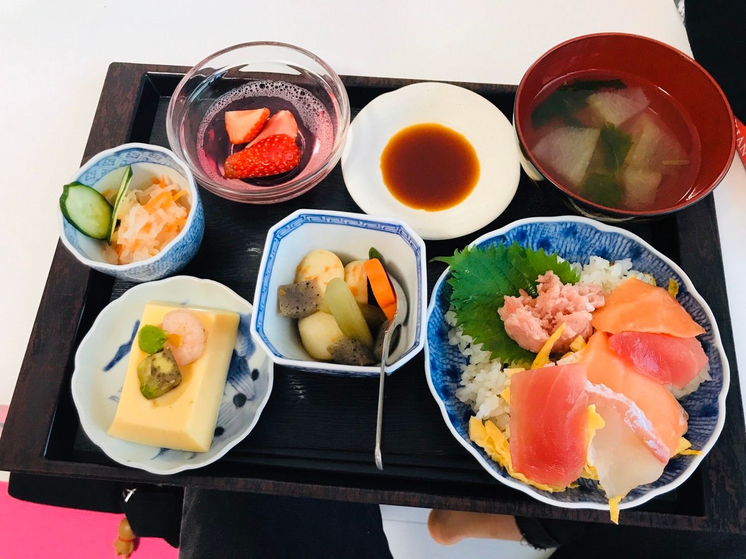ご飯に刺身をのせた海鮮丼と豆腐料理、煮物と野菜の小鉢、味噌汁、いちごのゼリーがトレーに並んでいる食事