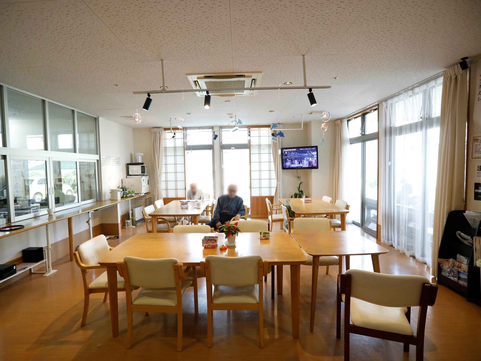 テーブルがいくつも並んでおり、入居者様が椅子に座り、くつろいでる。テレビが設置されており、映像が流れている。