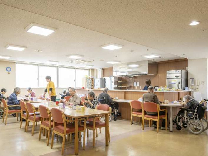 食堂には複数のダイニングテーブルがあり、入居者が食卓を囲んでいる。スタッフが一名見守りをしていて、キッチン内にも一名いる。