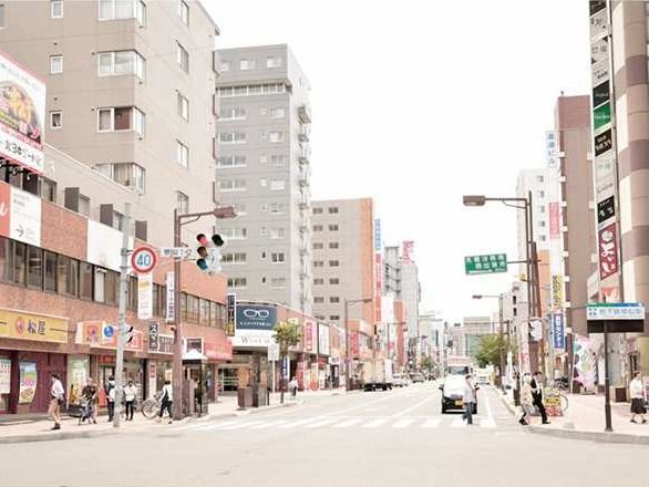 飲食店やビルが建ち並ぶにぎやかな街中の風景。道路にはバイクや車が通っていて、通行人も多い。