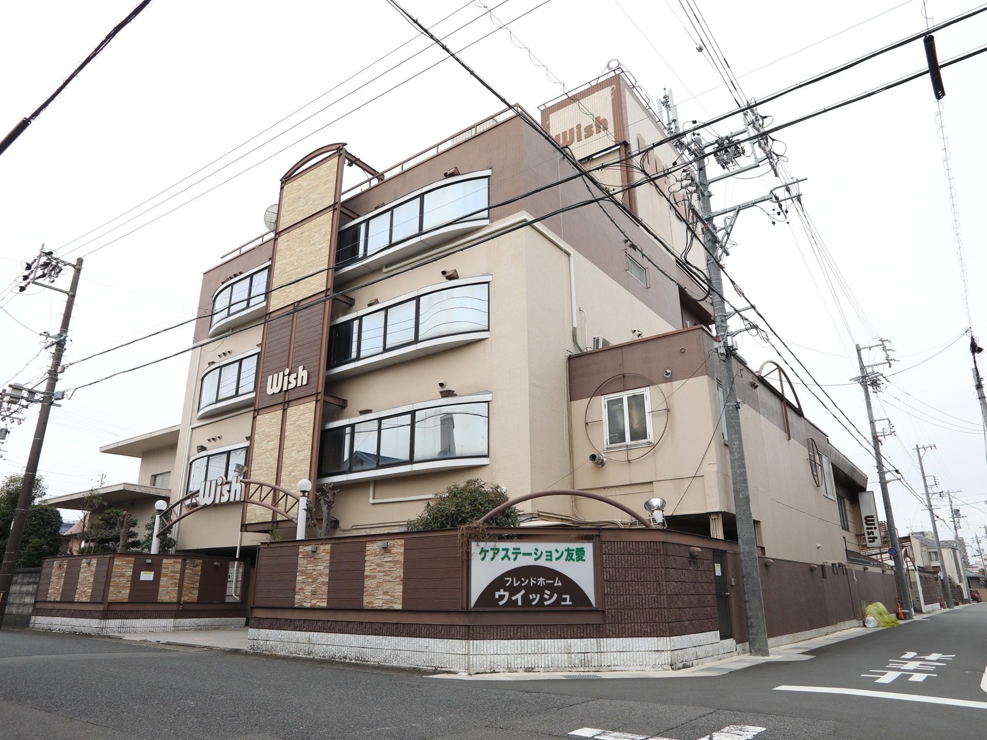 建物の周りの塀に施設名が書かれたプレートが設置されている。外壁は薄いブラウンと濃いブラウンで統一されていて、4階建てである。