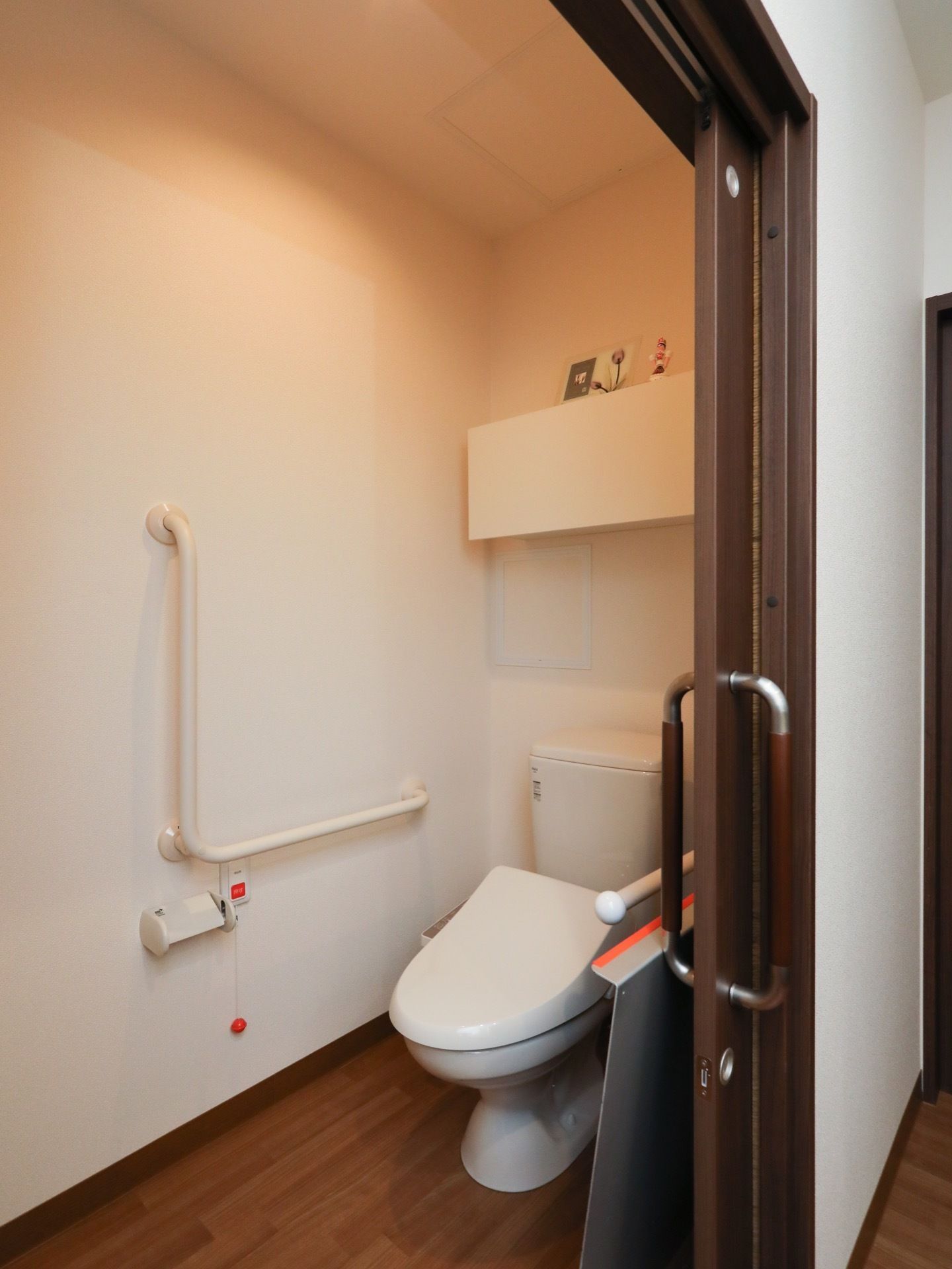 居室に備え付けの洋式トイレである。扉は引き戸式タイプ。壁にはＬ字型の手すり、ナースコールを設置している。