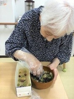 女性入居者がビニール手袋をして調理を行っている様子。木製のテーブルの上には具材が入れられた椀と牛乳パックがある。